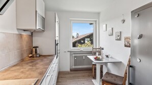 Una cucina compatta con mobili bianchi, una finestra con vista su una casa, un termosifone e un piccolo tavolo con una sedia.