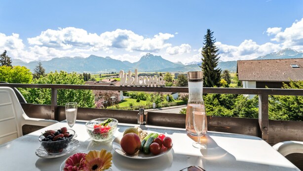 Un tavolo con un bicchiere d'acqua, un bicchiere di vino e una ciotola di frutta su un balcone con vista sulle montagne.