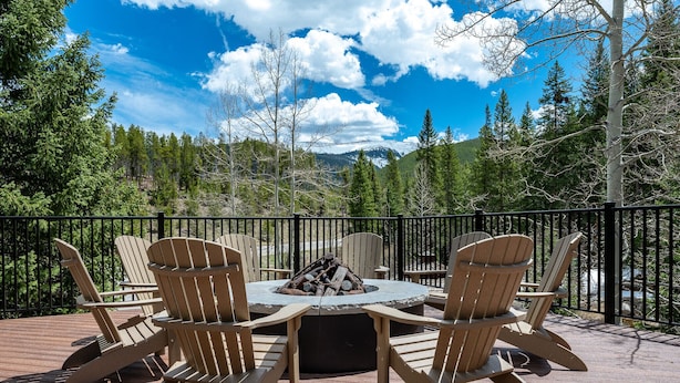 Una terraza con sillas Adirondack y un brasero central, rodeada por una barandilla metálica, con vistas a una cadena montañosa boscosa.