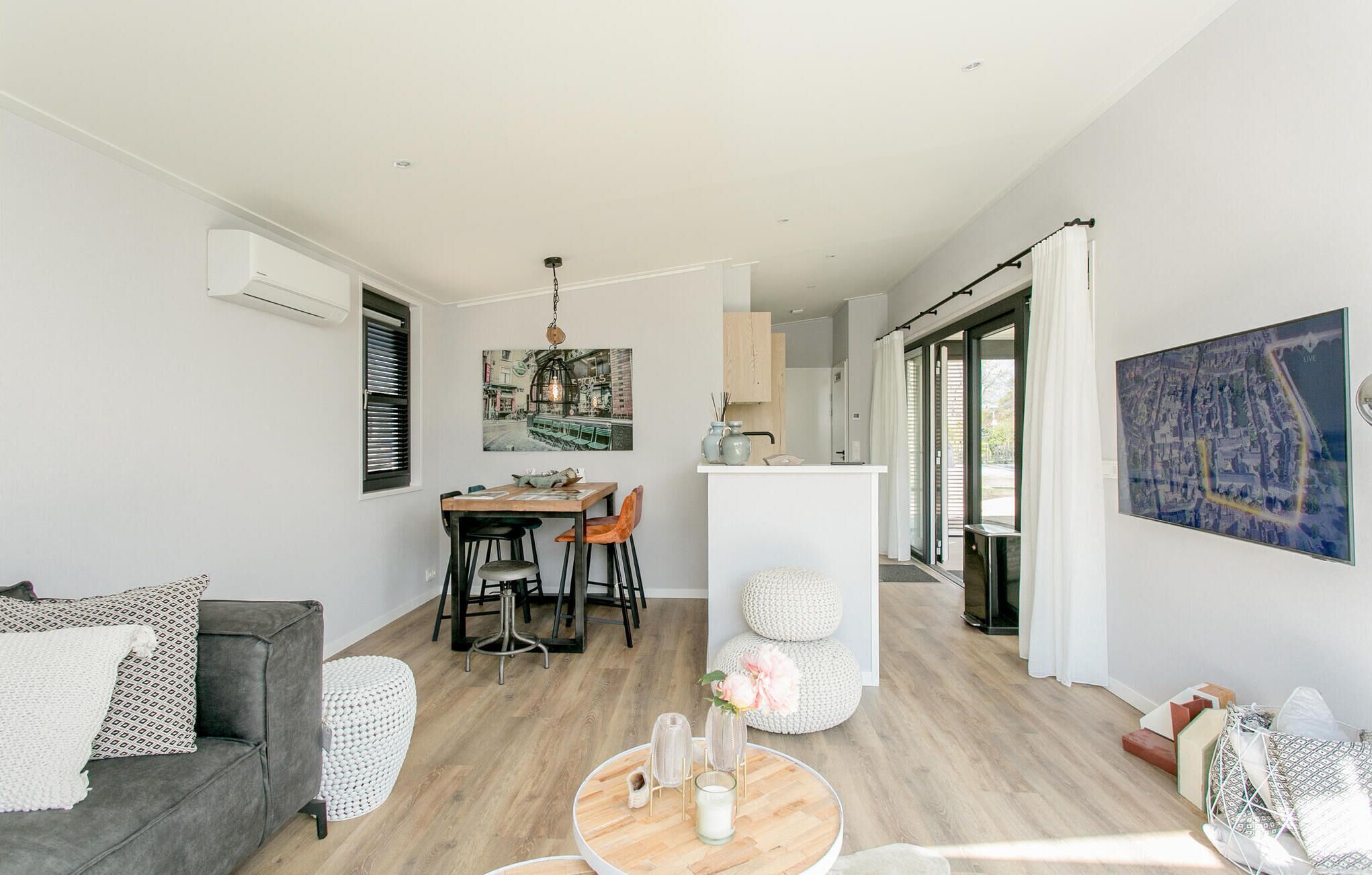A modern living room with a grey sofa, a dining area with a wooden table, and a kitchen with a white countertop.