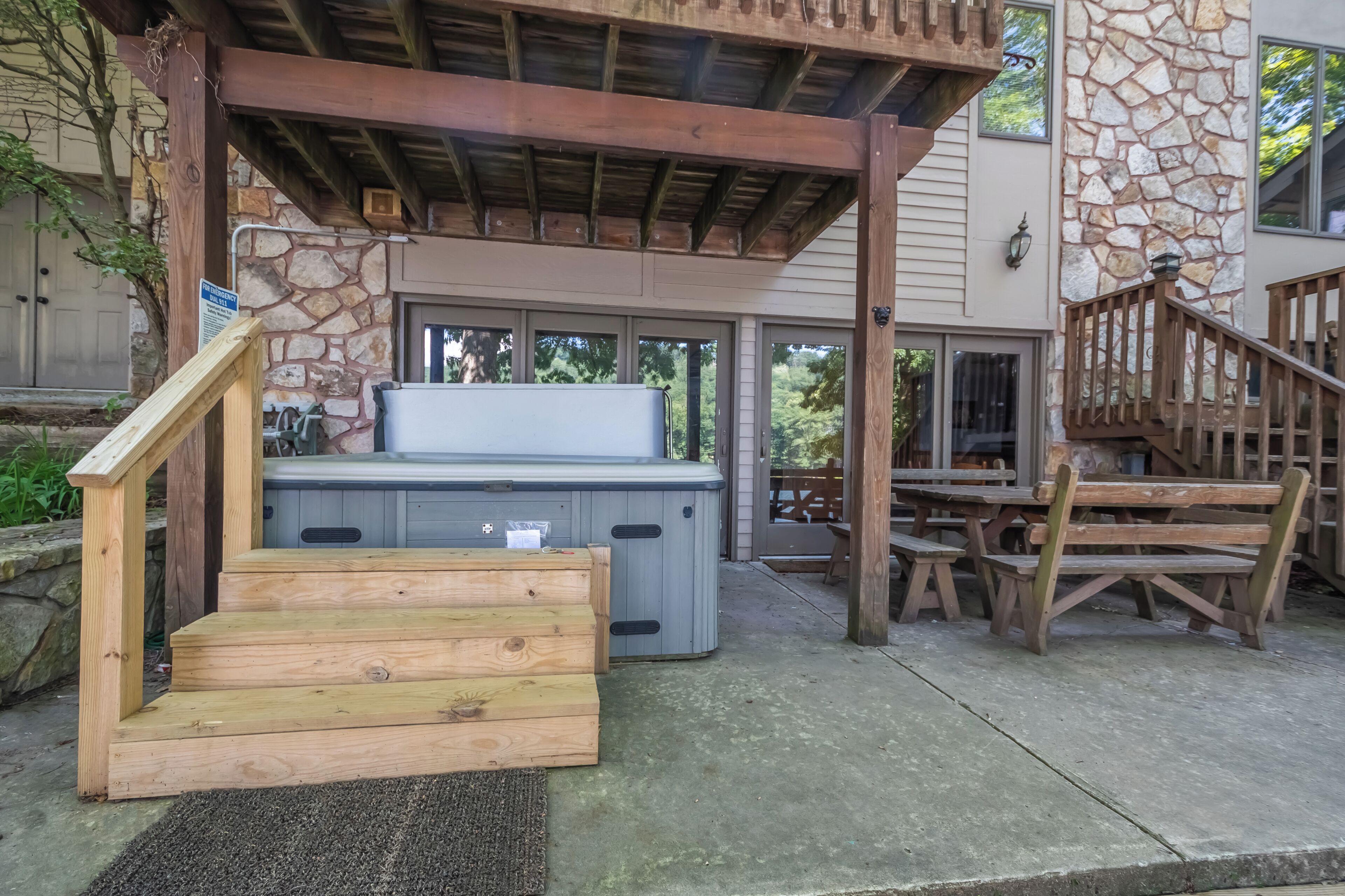 Un bain à remous sous une pergola en bois, avec un mur de pierres en arrière-plan, des marches en bois menant au bain, et un escalier en bois sur la droite.