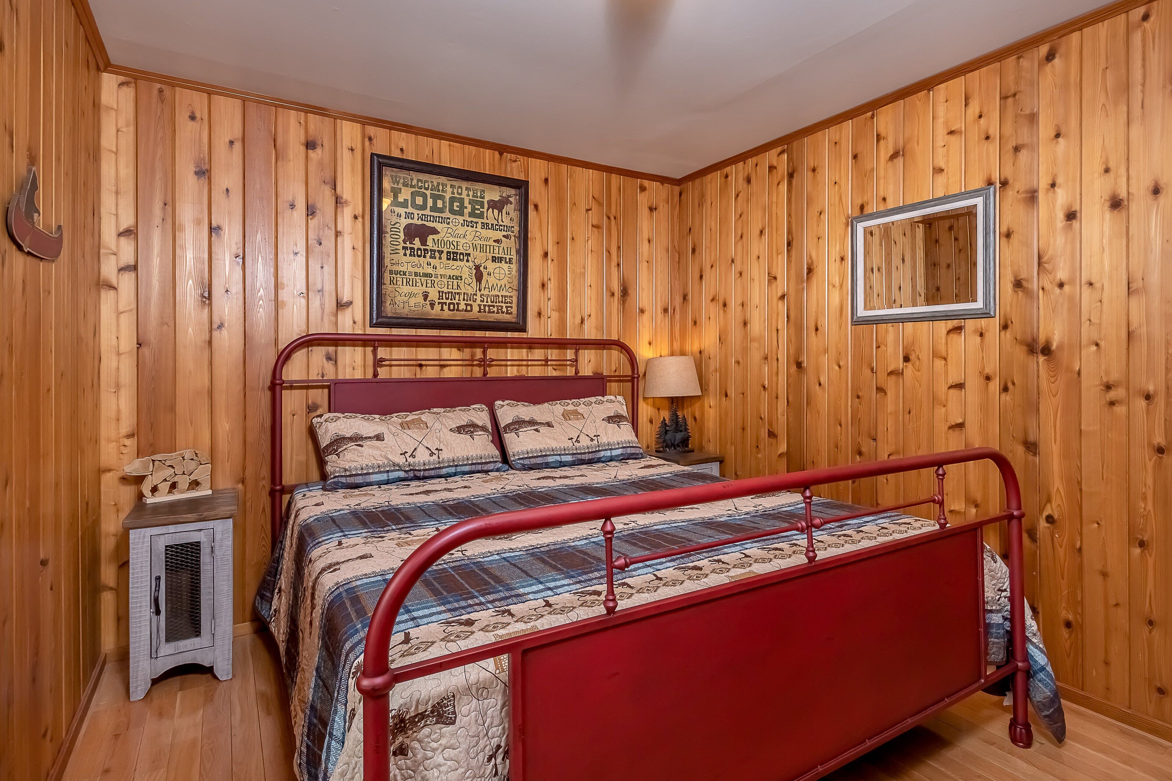 A wooden-paneled room with a red metal bed, a framed welcome sign, and a small table with a lamp.
