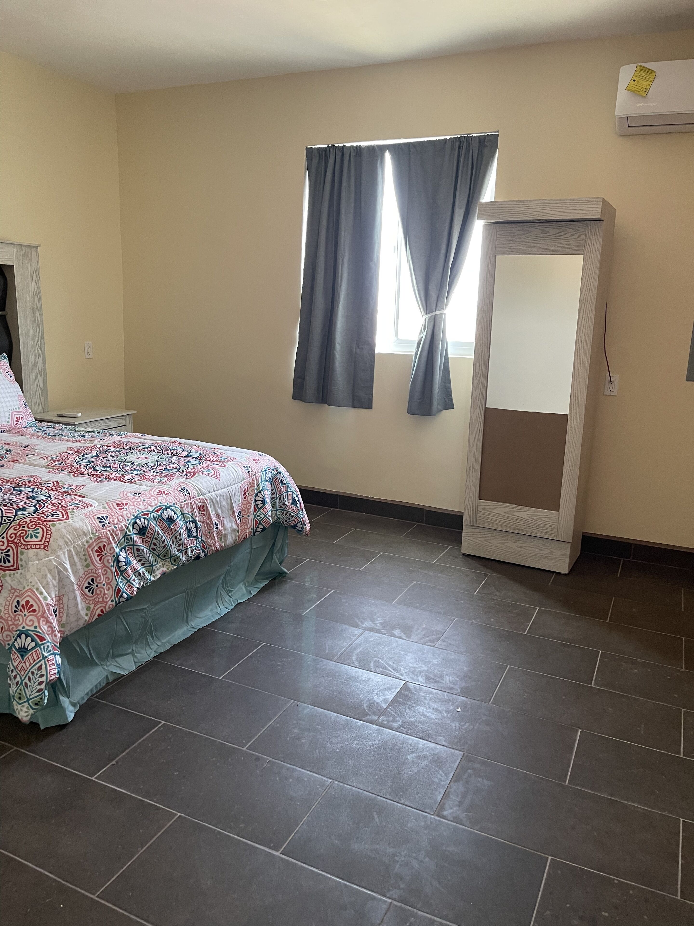 Une chambre à coucher avec un lit, une fenêtre avec des rideaux, un miroir et un sol carrelé.