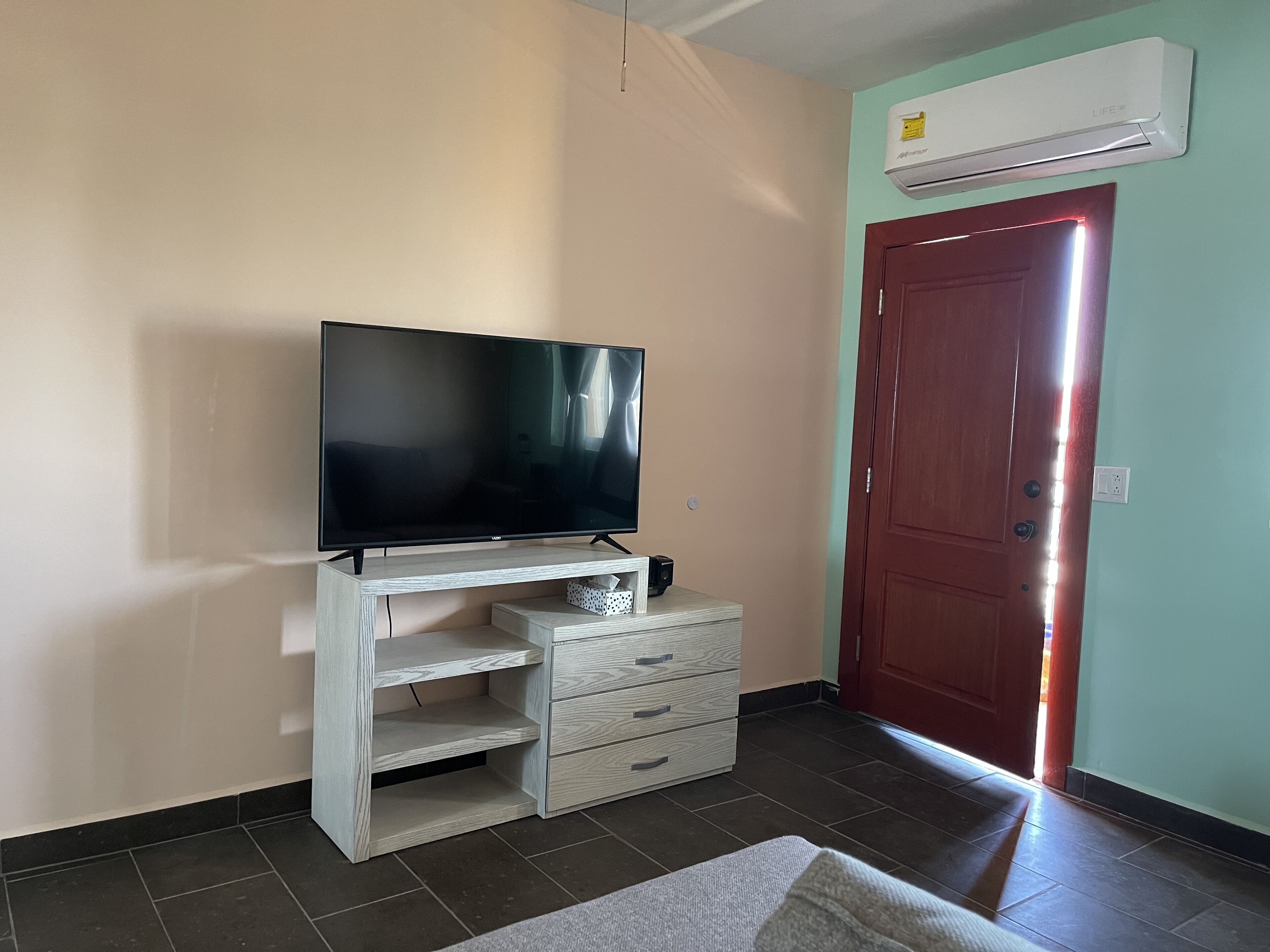 Habitación con una televisión de pantalla plana sobre un soporte de madera, una puerta roja y un equipo de aire acondicionado en la pared.