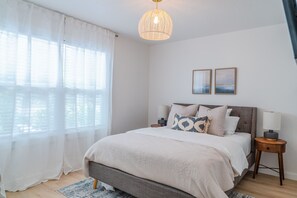 A bedroom with a bed, bedside lamps, a window with sheer curtains, and two wall-mounted pictures.