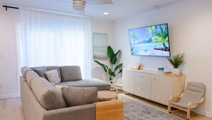 Ein modernes Wohnzimmer mit einem grauen Sofa, einem Holztischchen, einem Korbstuhl, einer Holzkabine und einem großen Fernseher an der Wand.