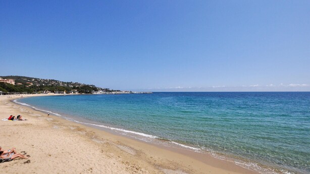 Ein Strand mit klarem, blauem Wasser, sandigem Ufer und Gebäuden im Hintergrund.