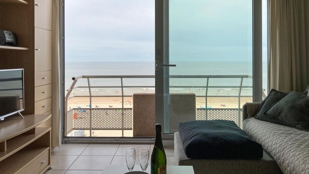 A room with a large sliding glass door leading to a beach view, a sofa, a TV on a stand, and a bottle of champagne. - Gorgeous apartment in Oostende (Oostende)