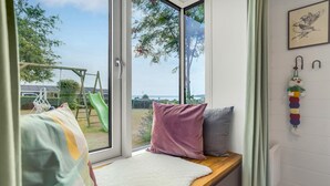 Een kamer met een groot raam, een houten bank met kussens en uitzicht op een speeltuin en bomen.