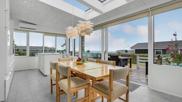 Een moderne keuken met een groot eiland, een eethoek met een houten tafel en stoelen, en grote ramen met uitzicht op zee en het omliggende landschap.
