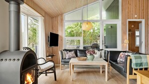 En hyggelig stue med brændeovn, sofa, sofabord og store vinduer med udsigt til grønne omgivelser.