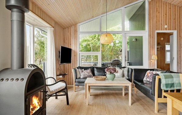 A cozy living room with a wood-burning stove, a sofa, a coffee table, and large windows offering a view of greenery outside.