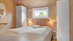 Et soveværelse med træloft, en seng med et mønstret tæppe, et vindue med udsigt til træer, og en hvid garderobe.