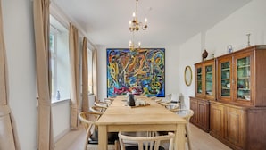 A dining room with a long wooden table, beige curtains, a large abstract painting, a chandelier, and a wooden cabinet with glass doors. - 6 bedroom gorgeous home in Kværndrup (Kværndrup)