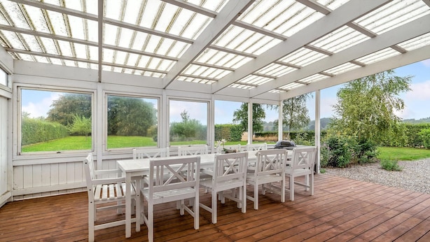 A covered patio with a wooden deck, white furniture, and large windows offering a view of a green landscape. - 6 bedroom gorgeous home in Kværndrup (Kværndrup)
