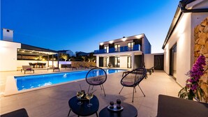 Un espace piscine extérieur moderne, doté de chaises longues et d’une table à manger, entouré d’un bâtiment à deux étages avec de grandes fenêtres et un mur en pierre.