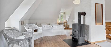 Un dormitorio acogedor en el ático, con cama, una silla de mimbre blanca, una mesita y un estufa de leña.