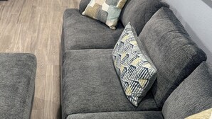 Moderni olohuone, jossa on harmaa sohva, sohvapöytä ja kuviollinen koristetyyny. Taustalla näkyy keittiö, jossa on valkoiset kaapit ja mikroaaltouuni.