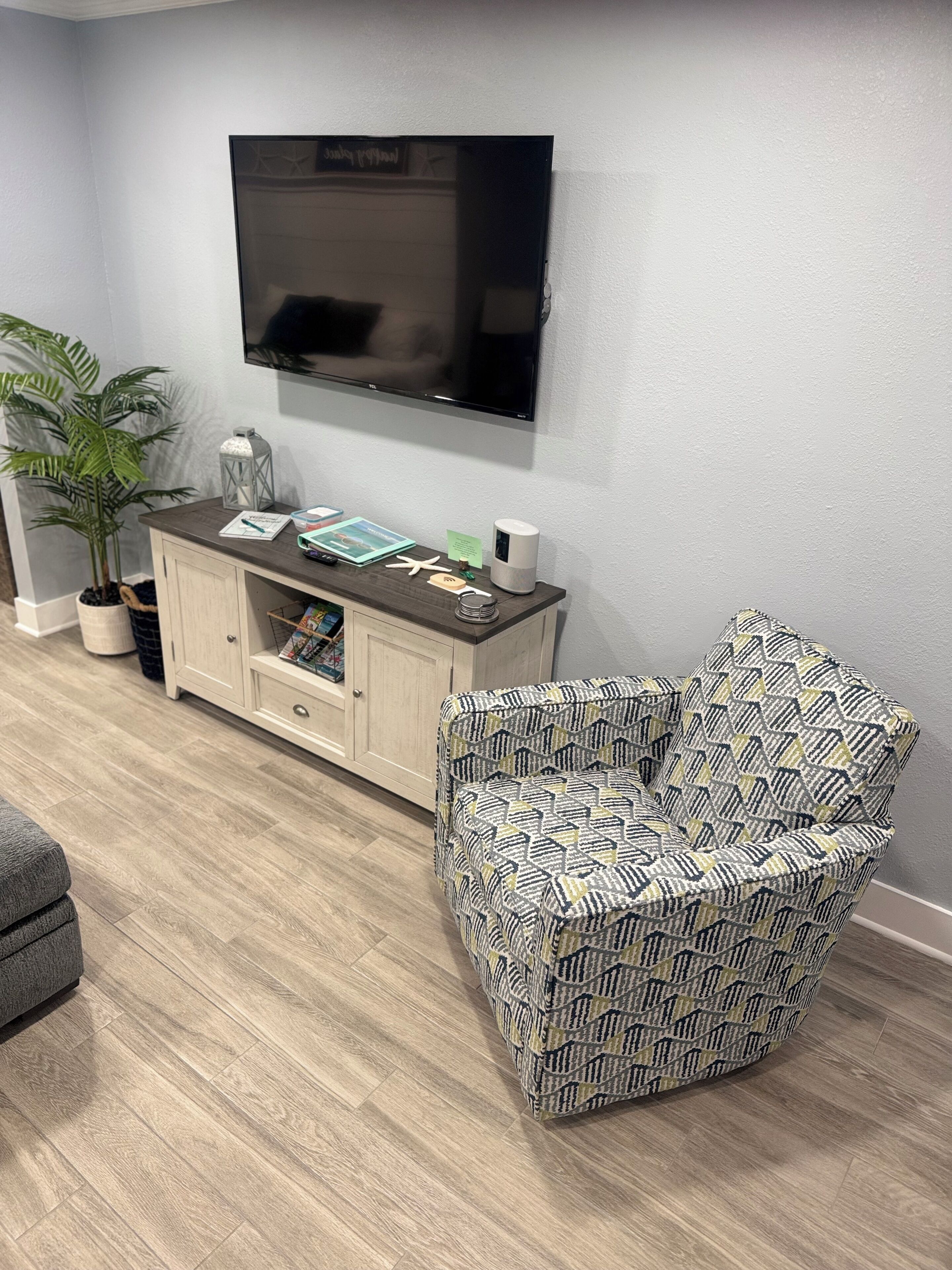 Een woonkamer met een aan de muur bevestigde televisie, een kast met tijdschriften, een gestreept fauteuil en een potplant.