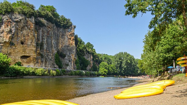 Ein Fluss mit gelben Kanus, die am Ufer liegen, ein felsiger Abhang und Bäume im Hintergrund.