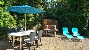 Eine Terrasse im Freien mit einem runden Tisch, vier Stühlen, einem blauen Sonnenschirm, einer Feuerstelle aus Ziegelsteinen und zwei türkisfarbenen Liegestühlen.