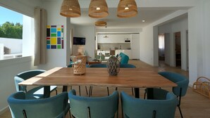 A modern dining area with a wooden table and blue chairs, a kitchen in the background, and a large window with curtains.