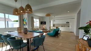 A spacious living area with a wooden dining table, teal chairs, and a sofa. There are wicker pendant lights, a TV, and a kitchen in the background.