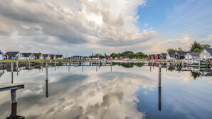 A calm waterfront with docked boats, a pier, and a row of colorful houses reflecting in the water.
