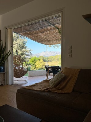 A living room with a sofa, a dining table, and chairs. There is a large window with a view of a mountain and greenery.
