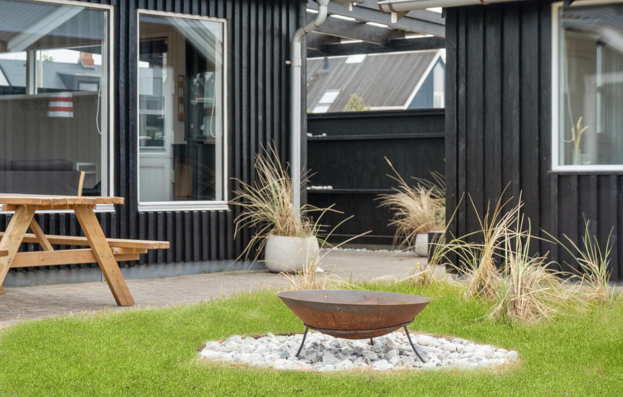 Un espace extérieur moderne avec un foyer en métal entouré de gravier et d’herbe, un banc en bois et de hautes herbes.