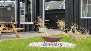 Un espace extérieur moderne avec un foyer en métal entouré de gravier et d’herbe, un banc en bois et de hautes herbes.