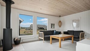 Un salon chaleureux avec une cheminée, deux canapés, une table basse et de grandes fenêtres donnant sur l’extérieur.