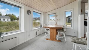 Une pièce avec un plafond en bois, des murs blancs, une table à manger en bois avec des chaises, de grandes fenêtres et une vue sur les maisons à l’extérieur.