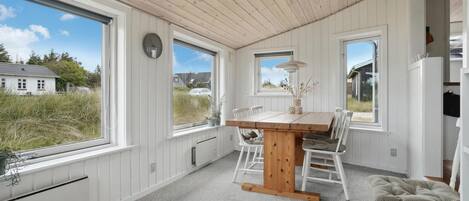 A room with a wooden ceiling, white walls, a wooden dining table with chairs, large windows, and a view of houses outside.