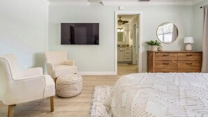 Une chambre à coucher comprenant un lit, une chaise, un pouf rond, une commode, un miroir et un téléviseur fixé au mur.