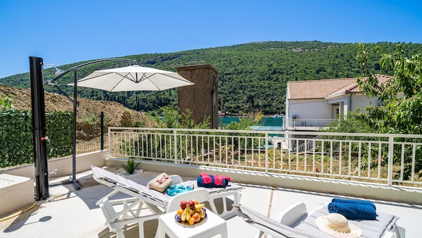 Un balcon avec des fauteuils de jardin blancs, une table garnie de fruits et une vue sur une colline et l’eau.