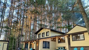 A two-story cabin with a covered patio area, outdoor seating, and a view of tall pine trees. - Fall Splendor! Cozy Cabin, Lake Views & Campfires! (Cadillac)