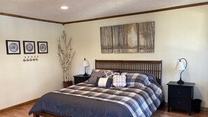A bedroom with a wooden bed, a nightstand with a lamp, a wall-mounted shelf, and a painting above the bed. - Fall Splendor! Cozy Cabin, Lake Views & Campfires! (Cadillac)
