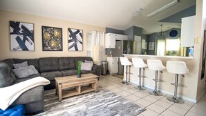 A modern living room with a grey sofa, a wooden coffee table, and a kitchen area with white bar stools and a refrigerator.