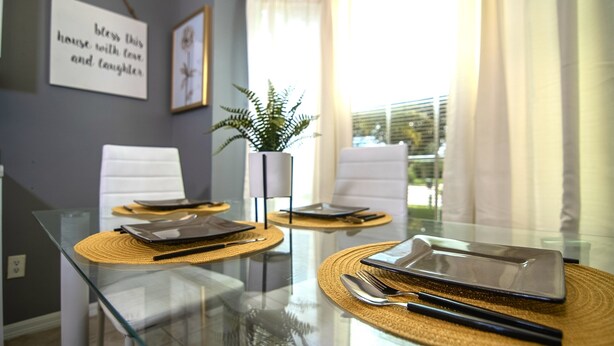 A dining table with placemats, cutlery, and a tablet. There are two chairs and a plant in the background.