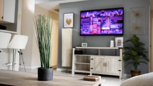 A modern living room with a wooden coffee table, a potted plant, a flat-screen TV mounted on the wall, and a minimalist cabinet.