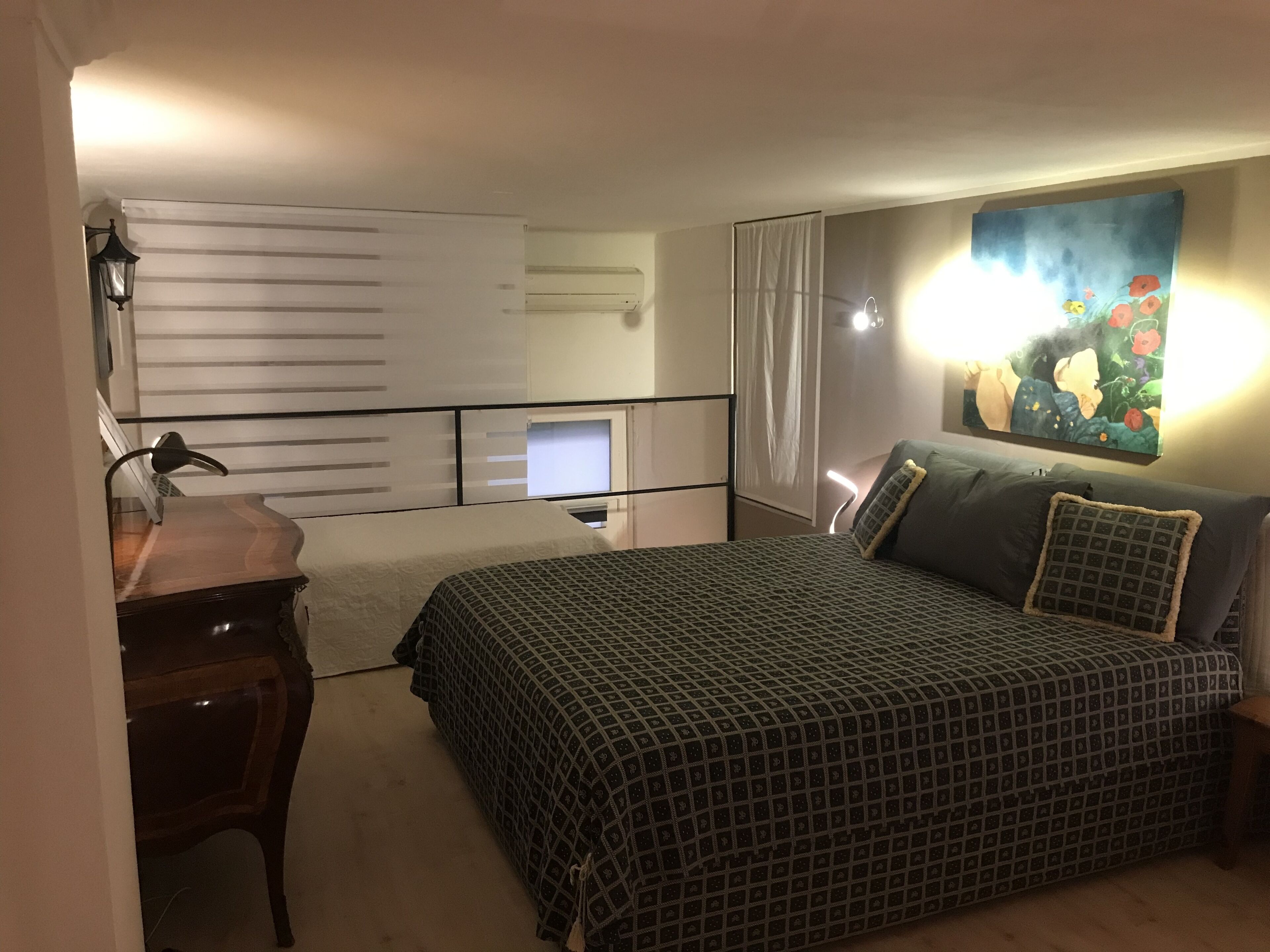 A bedroom with a bed, a desk, a chair, a painting on the wall, and a window with curtains.