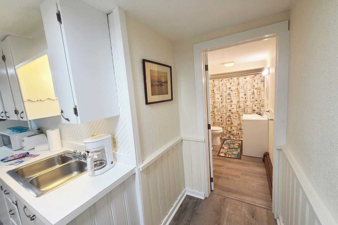 A narrow hallway leading to a bathroom with nautical-themed wallpaper, a sink with a coffee maker, and a framed picture on the wall.