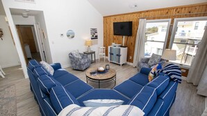 A modern living room with a large blue sectional sofa, a small round coffee table, and a TV mounted on the wall. - Semi-Oceanfront, Private Pool, Hot Tub, Close to beach! WL1066, Schemering Seas (Corolla)