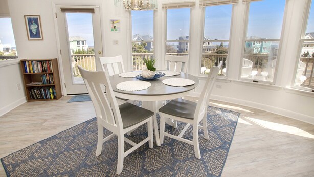 A dining area with a round table, white chairs, a chandelier, and a view through large windows. - Semi-Oceanfront, Private Pool, Hot Tub, Close to beach! WL1066, Schemering Seas (Corolla)