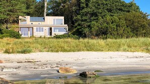Une maison moderne à deux étages, dotée de grandes fenêtres, située sur une plage de sable fin, avec une eau cristalline et des rochers.