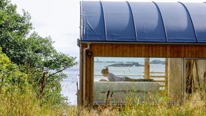 Une cabane en bois dotée d’une grande fenêtre donnant sur une étendue d’eau et des affleurements rocheux.