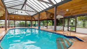 Una piscina interior con techo de madera y paredes de vidrio, rodeada por un suelo de baldosas y algunas sillas verdes.