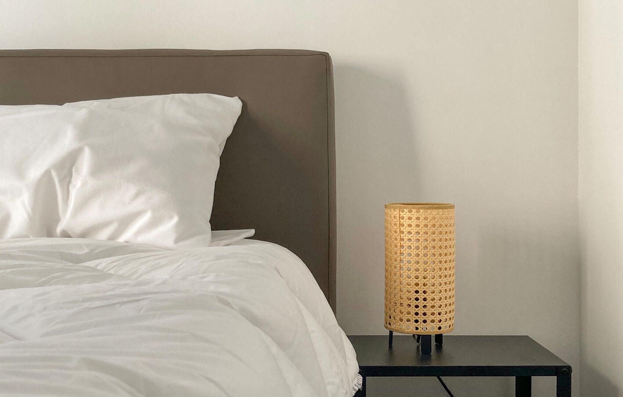 A bed with white bedding and a grey headboard, a black nightstand with a woven lamp, and a plain wall.