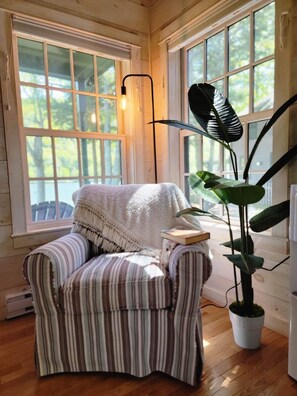 A cozy room with a striped armchair, a potted plant, and a floor lamp. - Cozy lakefront cottage w/seasonal hot tub. Mountain views, minutes to town/ocean (Camden)
